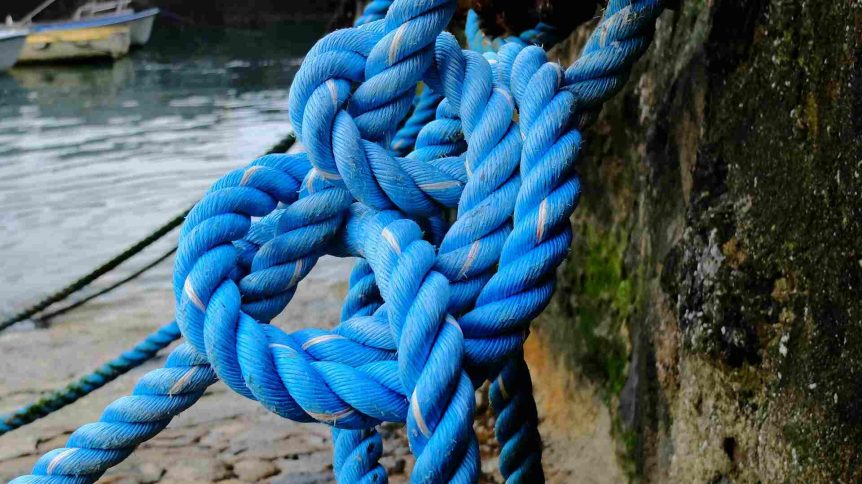 Close-up of a thick blue rope tied in complex knots, hanging near a weathered stone wall by the water.